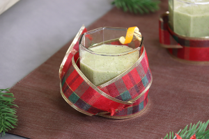 green smoothie in clear glass with orange peel garnish. Red plaid ribbon curled around glass.