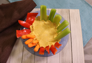 hummus in blue bowl with sliced red peppers, carrots, and celery on a cream colored board with a brown placemat