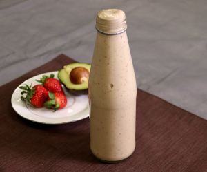 smoothie in tall glass bottle in front of white plate with fresh strawberries and half an avocado. Brown placemat on grey background.