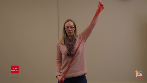 A woman in a pink sweater demonstrates how to stretch a red stretch band.