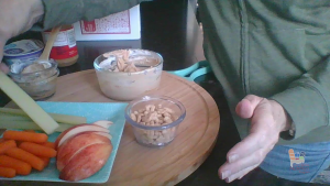 Female hands stand next to a plate of apples, carrots, and celery next to a bowl of cereal with yogurt dip in the background.