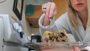 blonde woman in white bathrobe holds a plate with a blueberry muffin and cuts it with a fork