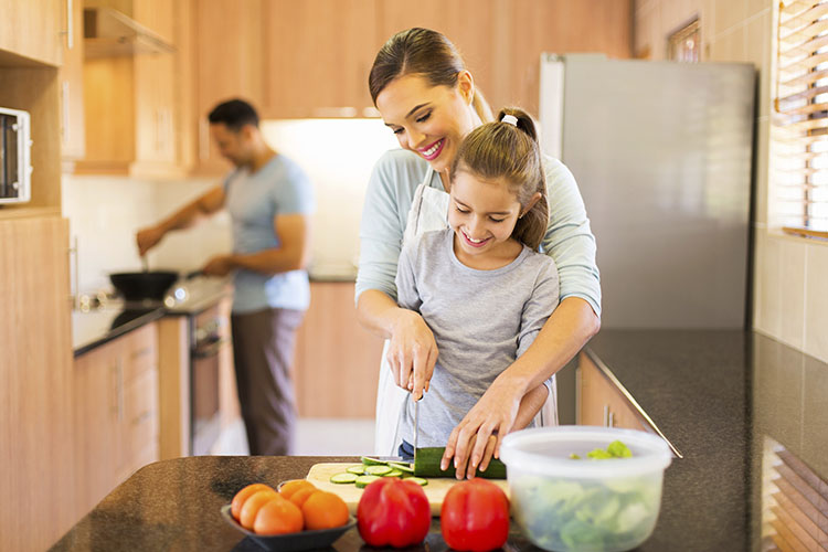 Mother and daughter cut a cucumber together while the dad stirs something on the stove in the background