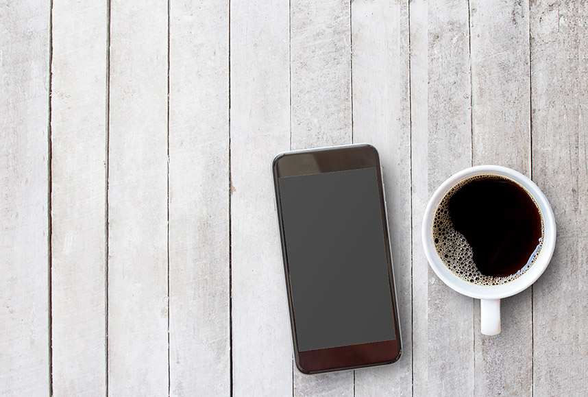 Smart phone and cup of coffee in a white cup on a white wooden surface.