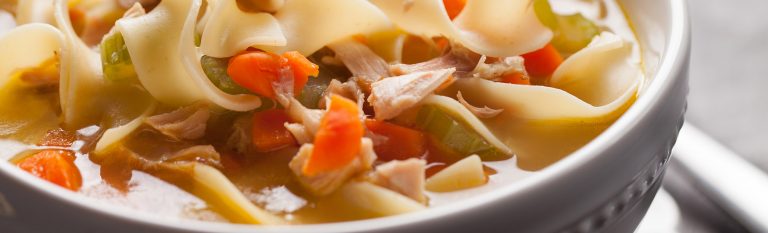white bowl of soup with chicken or turkey, carrots, and noodles.