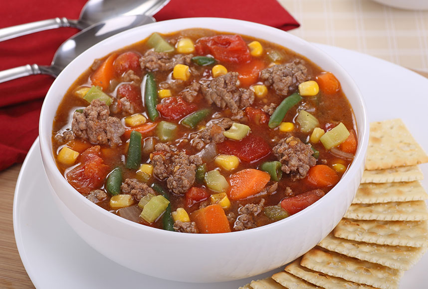 White bowl of beef and vegetable soup with crackers.