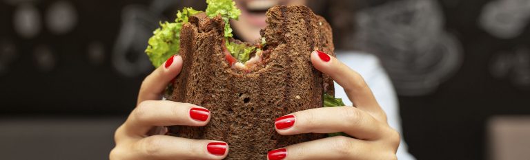 Whole-grain sandwich with bite taken out of it, held up by a woman smiling.