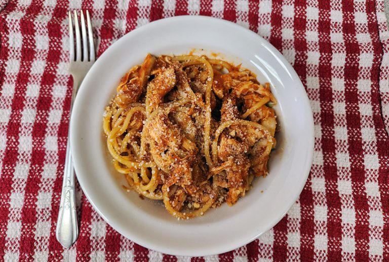White bowl of pasta with tomato sauce and chicken on a red and white checkered background.