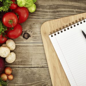 Paper and pen for planning set on cutting board surrounded by colorful vegetables set on a wooden table.