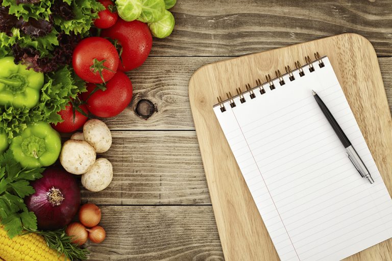 Paper and pen for planning set on cutting board surrounded by colorful vegetables set on a wooden table.