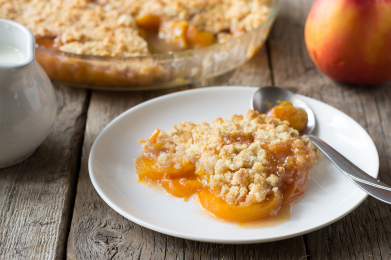 Hearty scoop of fruit crisp on a plate surrounded by peaches and cream, with more fruit crisp in dish in the background.