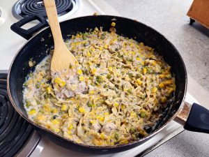 Skillet filled with noodles, fish, peas, and corn on a stove with a wooden spoon