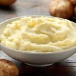 White bowl of creamy mashed potatoes with raw potatoes on the counter top