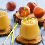 two glasses of peach smoothies with whole peaches in the background